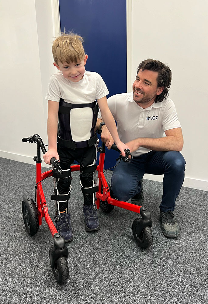 Patient with Reciprocating Gait Orthosis standing with his walker next to clinician Sam Walmsley, who made it for him
