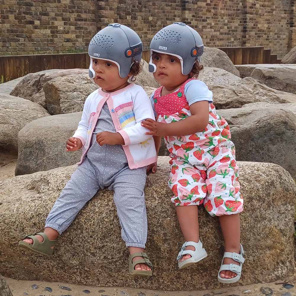 Identical twins with craniosynostosis during their cranial remoulding helmet therapy journey sitting on the beach wearing helmets
