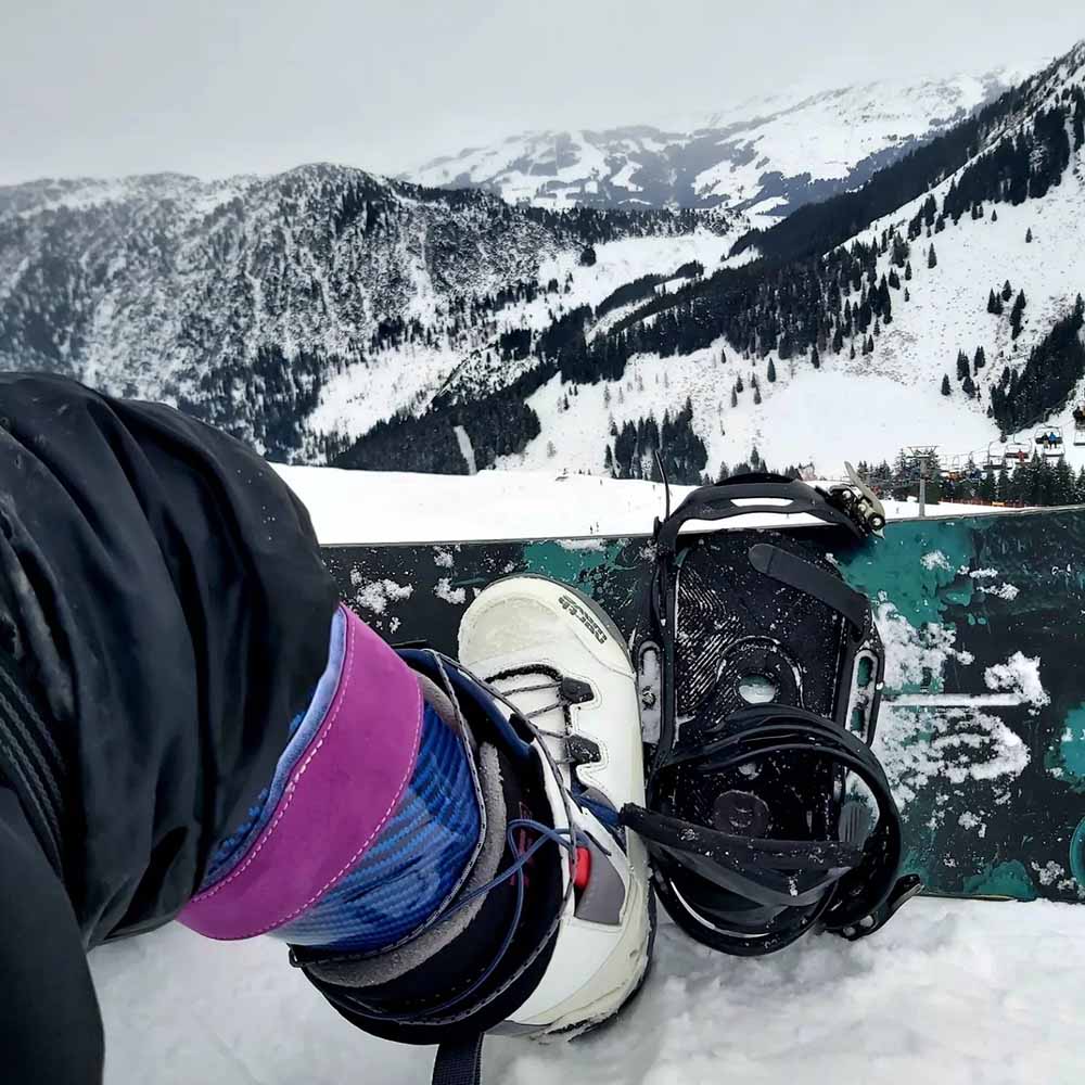 Close-up of a snowboarder’s boot and orthosis secured into a snowboard binding, with a snowy mountain landscape in the background