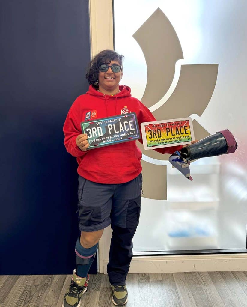 Nina Sparks smiling and holding two third-place awards while wearing her snowboarding gear and a custom orthosis on her left leg