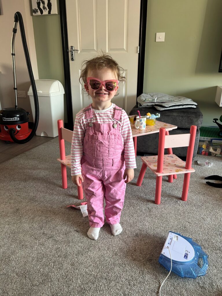 Lexie smiling in pink overalls and sunglasses, standing in a playroom after plagiocephaly treatment at LOC